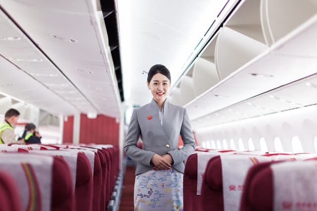 ลูกเรือ Hainan Airlines ในชุดยูนิฟอร์มสีเทา ยืนยิ้มในห้องโดยสาร